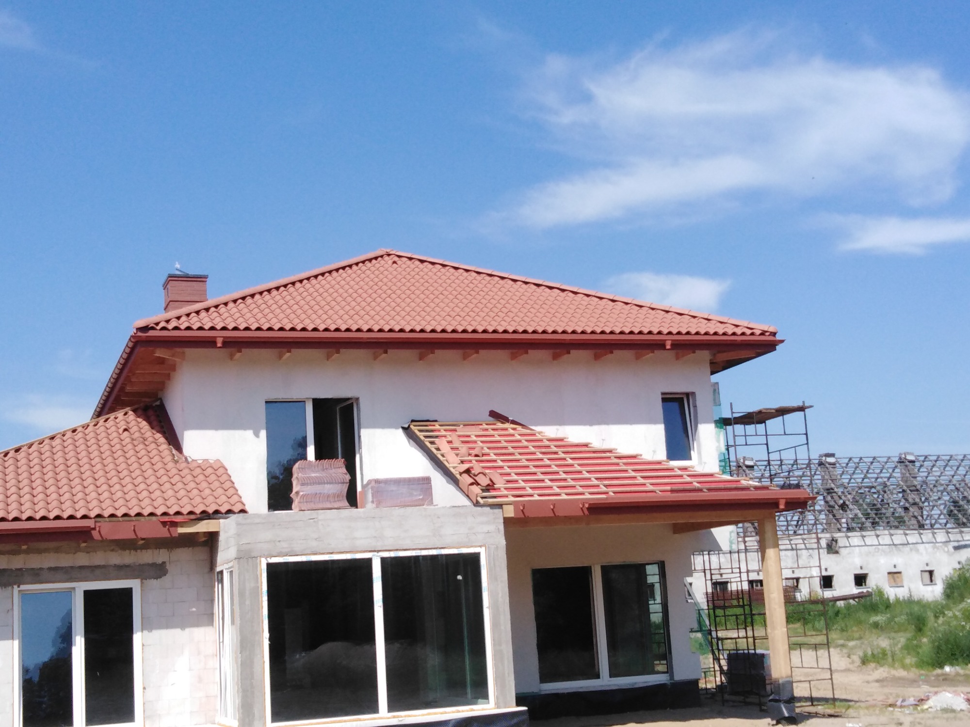 Dom jednorodzinny w trakcie krycia dachu czerwoną dachówką ceramiczną, widoczne rusztowanie i przygotowane dachówki, na kominie siedzi gołąb.