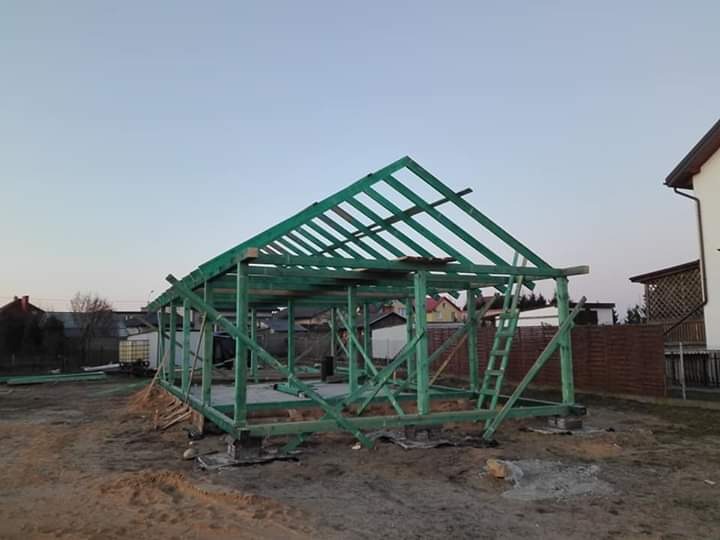 Konstrukcja drewniana dachu w trakcie budowy, zielona impregnacja, widoczna drabina oparta o szkielet, w tle zabudowa jednorodzinna.