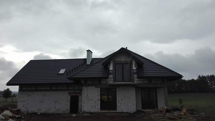 Dom w stanie surowym zamkniętym z czarnym dachem z dachówki ceramicznej, widoczny komin i okno dachowe, szare niebo w tle.