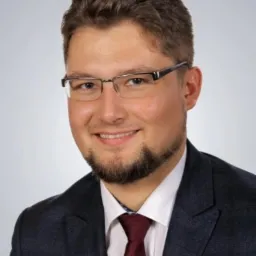 Portret uśmiechniętego mężczyzny w okularach, garniturze i krawacie na jasnym tle.