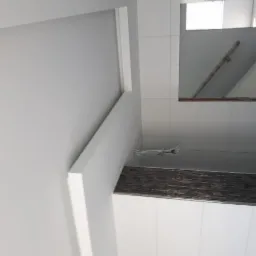 Wykończone wnętrze z białą ścianą, listwami dekoracyjnymi i fragmentem sufitu z widocznymi przewodami elektrycznymi, odbite w lustrze.