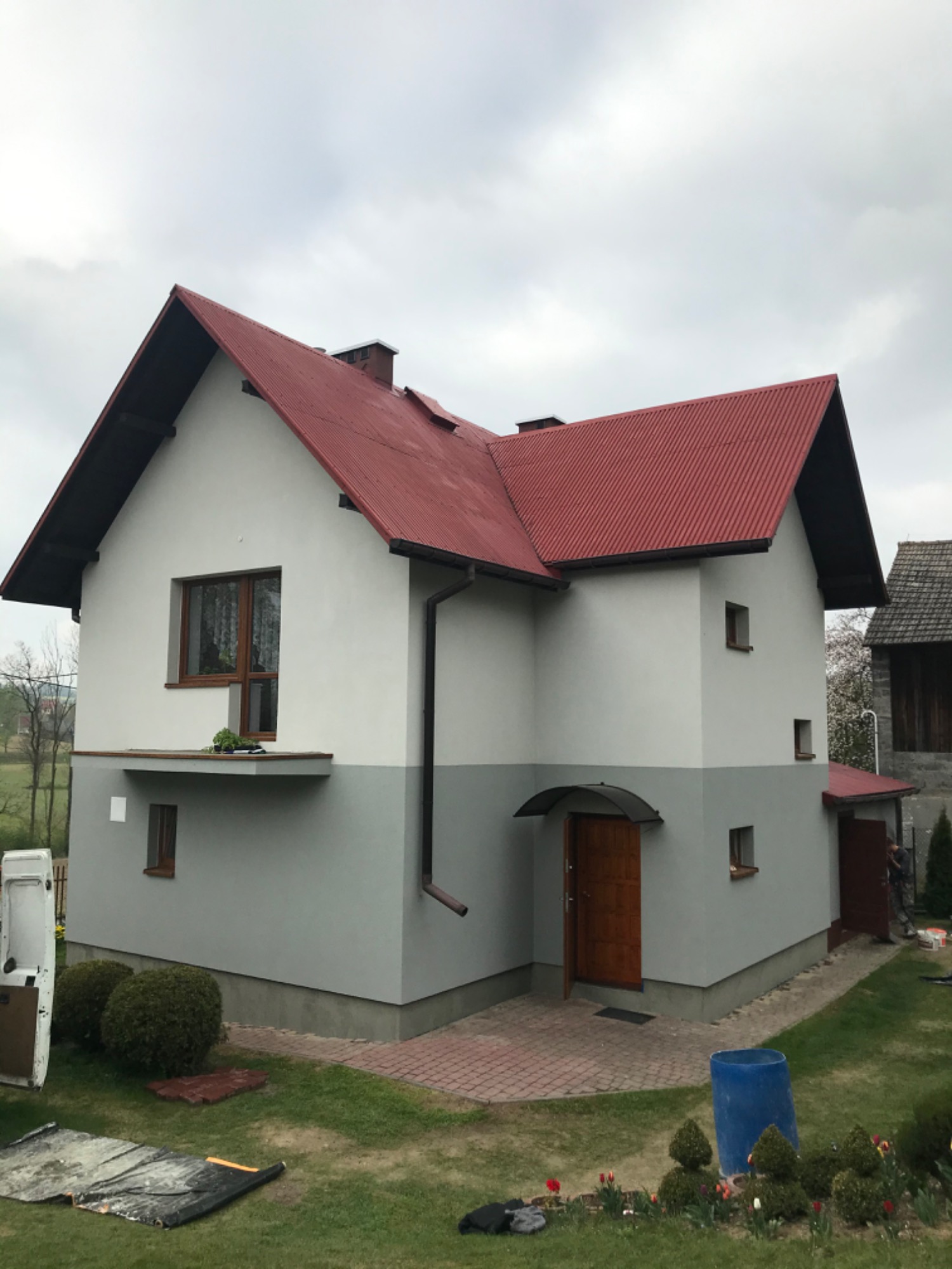 Dwukondygnacyjny dom z czerwoną blachodachówką i elewacją w dwóch odcieniach szarości, z widocznymi elementami wykończeniowymi, takimi jak rynny i parapety, na tle zachmurzonego nieba.