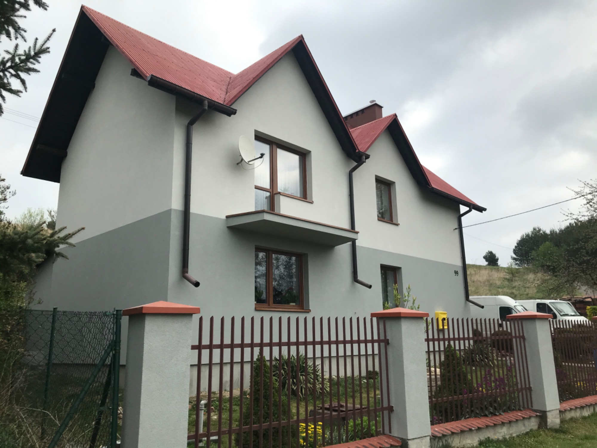 Dwukondygnacyjny dom z dwukolorową elewacją (szaro-białą), czerwonym dachem i brązowymi oknami, widziany zza ogrodzenia z metalowymi sztachetami i betonowymi słupkami.