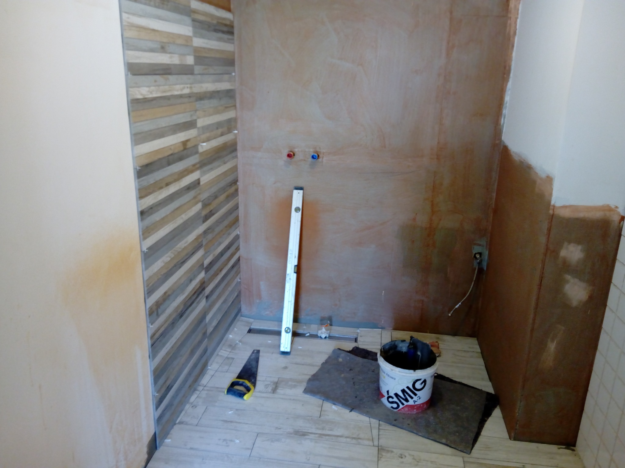 Niedokończona łazienka w trakcie układania płytek: ściana z poziomymi panelami imitującymi drewno, surowa ściana z wystającymi rurami, poziomica oparta o ścianę, wiadro z klejem SMIG i piła ręczna...