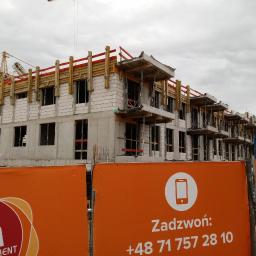 RV-Construction - Wielopiętrowy budynek w stanie surowym, z widocznym rusztowaniem i żurawiem w tle, częściowo zasłonięty pomarańczowym banerem reklamowym z numerem telefonu.