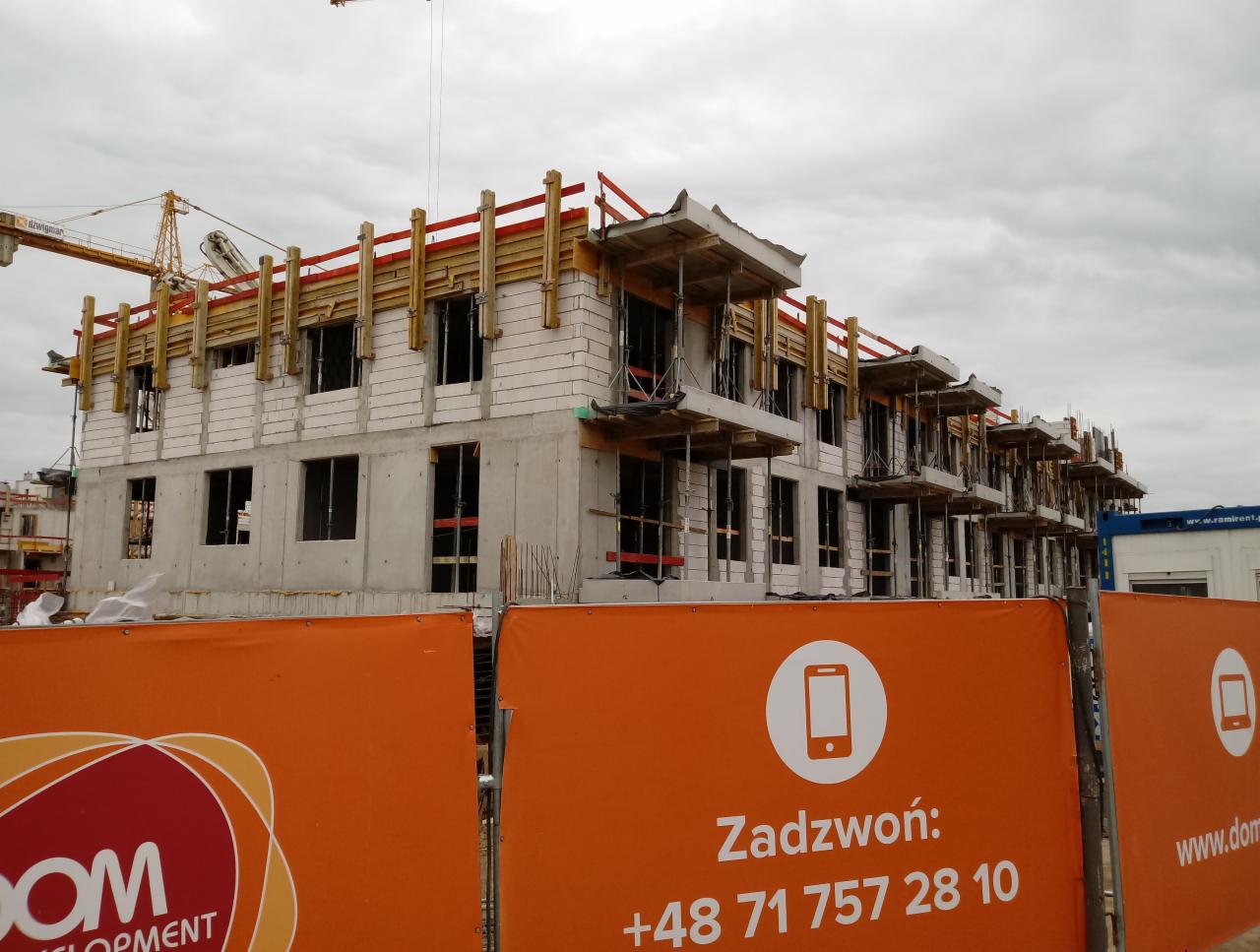Wielopiętrowy budynek w stanie surowym, z widocznym rusztowaniem i żurawiem w tle, częściowo zasłonięty pomarańczowym banerem reklamowym z numerem telefonu.