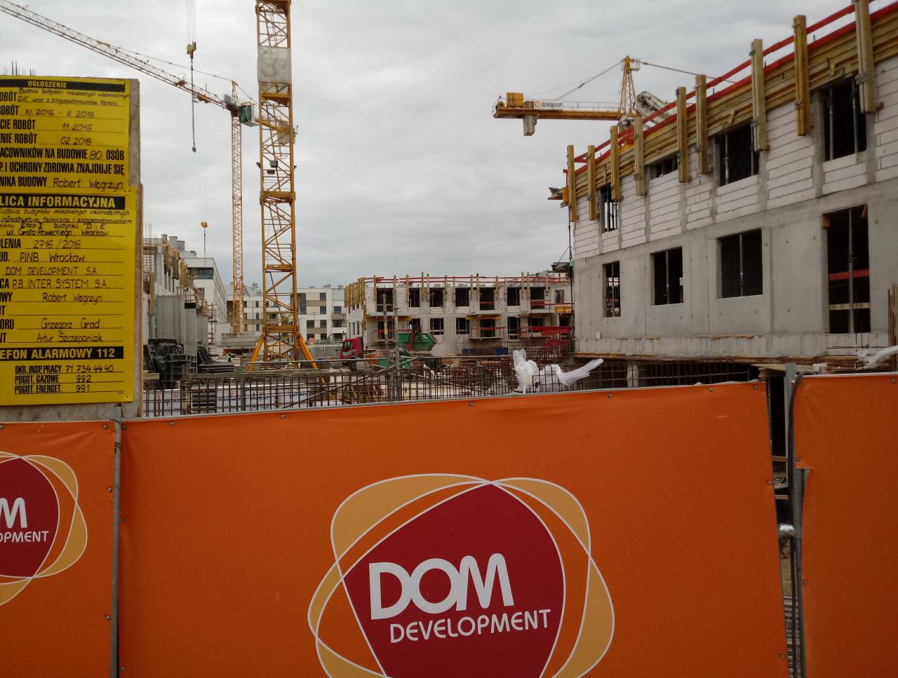 W budowie wielorodzinny budynek mieszkalny z widocznymi blokami z betonu komórkowego, rusztowaniami i dwoma żurawiami budowlanymi na tle pochmurnego nieba, zabezpieczony pomarańczową siatką z logo...