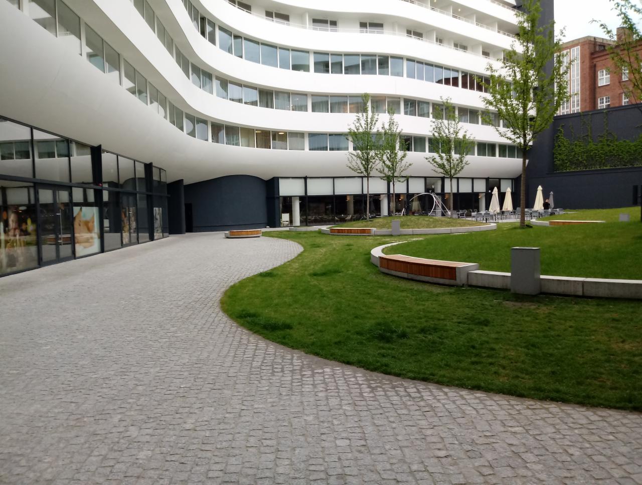 Kaskadowa ścieżka z szarej kostki prowadząca do nowoczesnego budynku z zaokrąglonymi balkonami, obok trawnik z drewnianymi ławkami i parasolami.