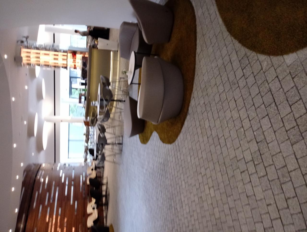 Nowoczesne wnętrze kawiarni lub restauracji z jasną kostką brukową, minimalistycznymi krzesłami barowymi i dekoracyjną ścianą z drewnianymi elementami.