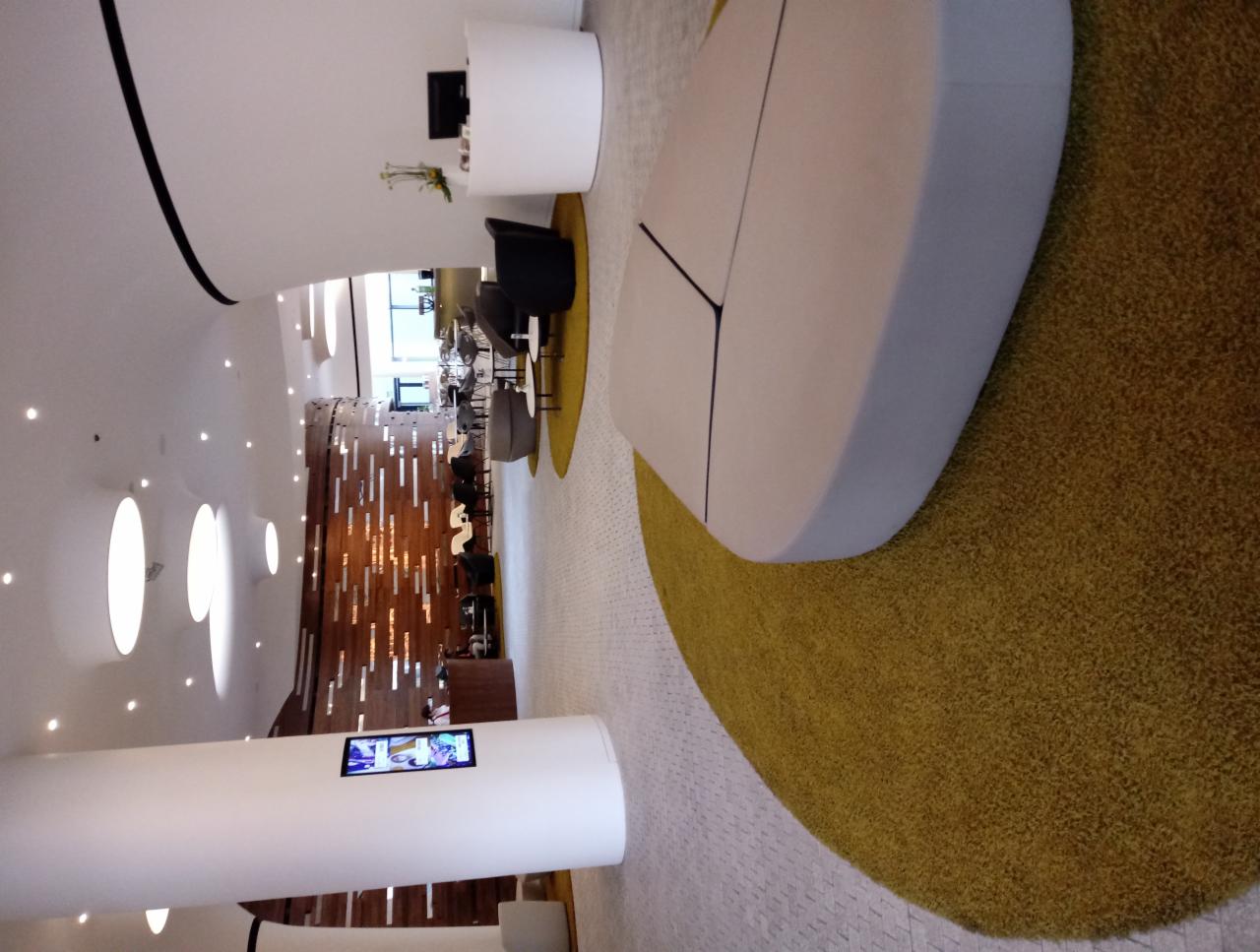 Nowoczesne wnętrze biurowe z białymi kolumnami, designerskimi meblami wypoczynkowymi i dekoracyjną ścianą z drewnianymi elementami, oświetlone punktowymi lampami sufitowymi.