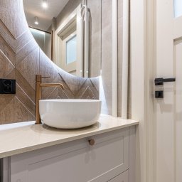 DD Design - Nowoczesna łazienka z okrągłym, podświetlanym lustrem, białą umywalką nablatową, złotą baterią, drewnianą ścianą w jodełkę i białymi grzejnikami dekoracyjnymi.