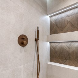 DD Design - Nowoczesna kabina prysznicowa z beżowymi płytkami, dekoracyjną ścianą z drewnianym wzorem chevron, miedzianą armaturą i subtelnym oświetleniem LED.
