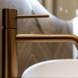 Miedziana bateria umywalkowa z prostą wylewką i uchwytem, na tle ściany z wzorem w jodełkę i fragmentu białej umywalki.