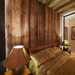 DD Design - Rustykalna sypialnia z drewnianymi ścianami i sufitem, etniczna pościel w odcieniach brązu i złota, lampka nocna z abażurem w kolorze terakoty, nóż leżący na drewnianym stoliku nocnym.