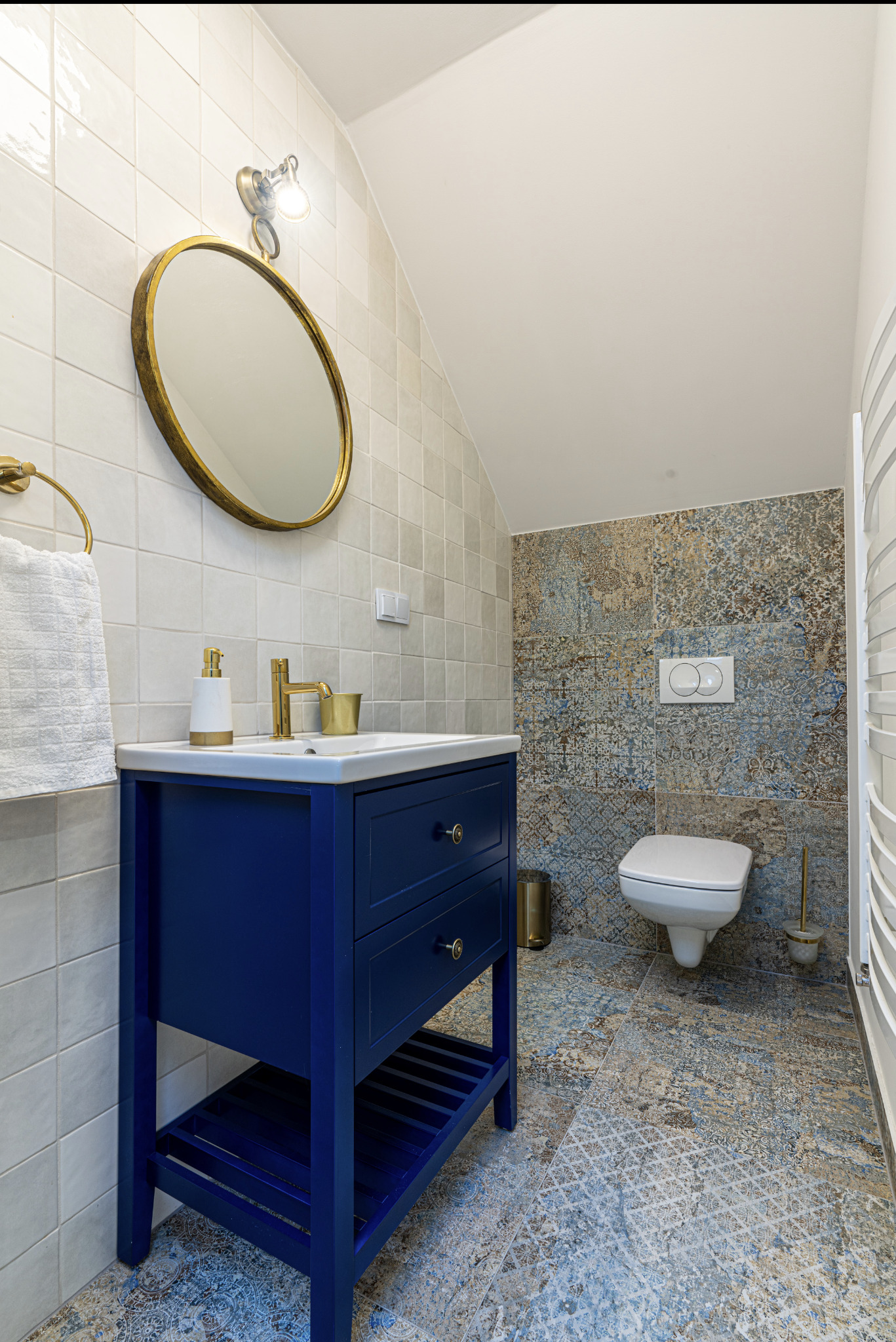 Niebieska szafka umywalkowa z owalnym lustrem w złotej ramie, złota armatura i mozaika na ścianie w nowoczesnej łazience na poddaszu.