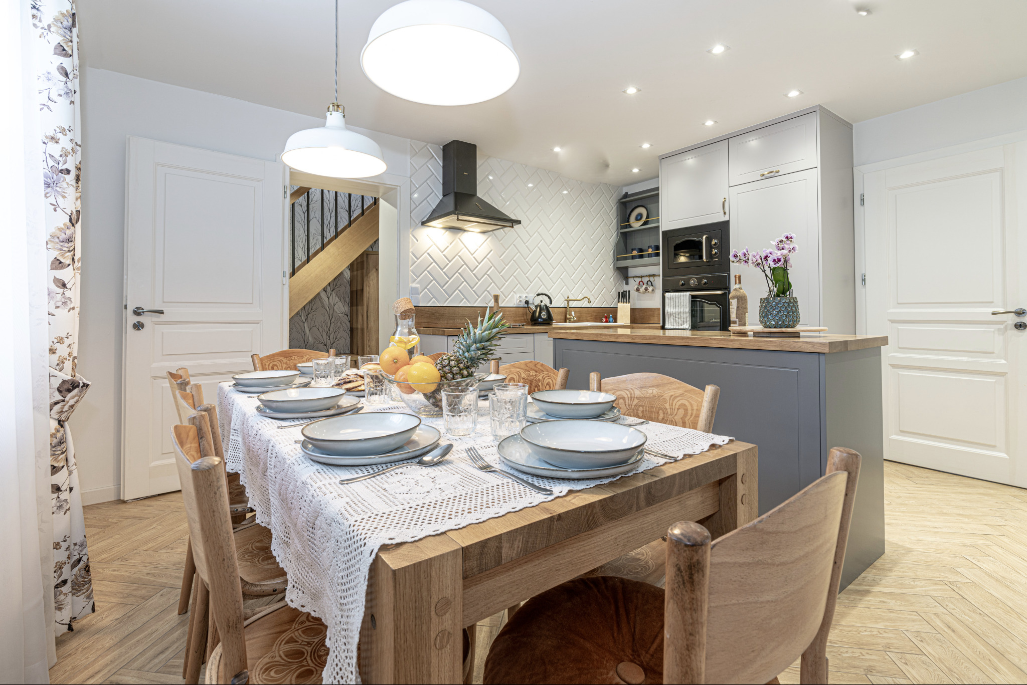 Jasna jadalnia z drewnianym stołem nakrytym białą koronkową serwetą, szare talerze, owoce w szklanej misie, w tle otwarta kuchnia z białymi płytkami ułożonymi w jodełkę i szarymi szafkami.