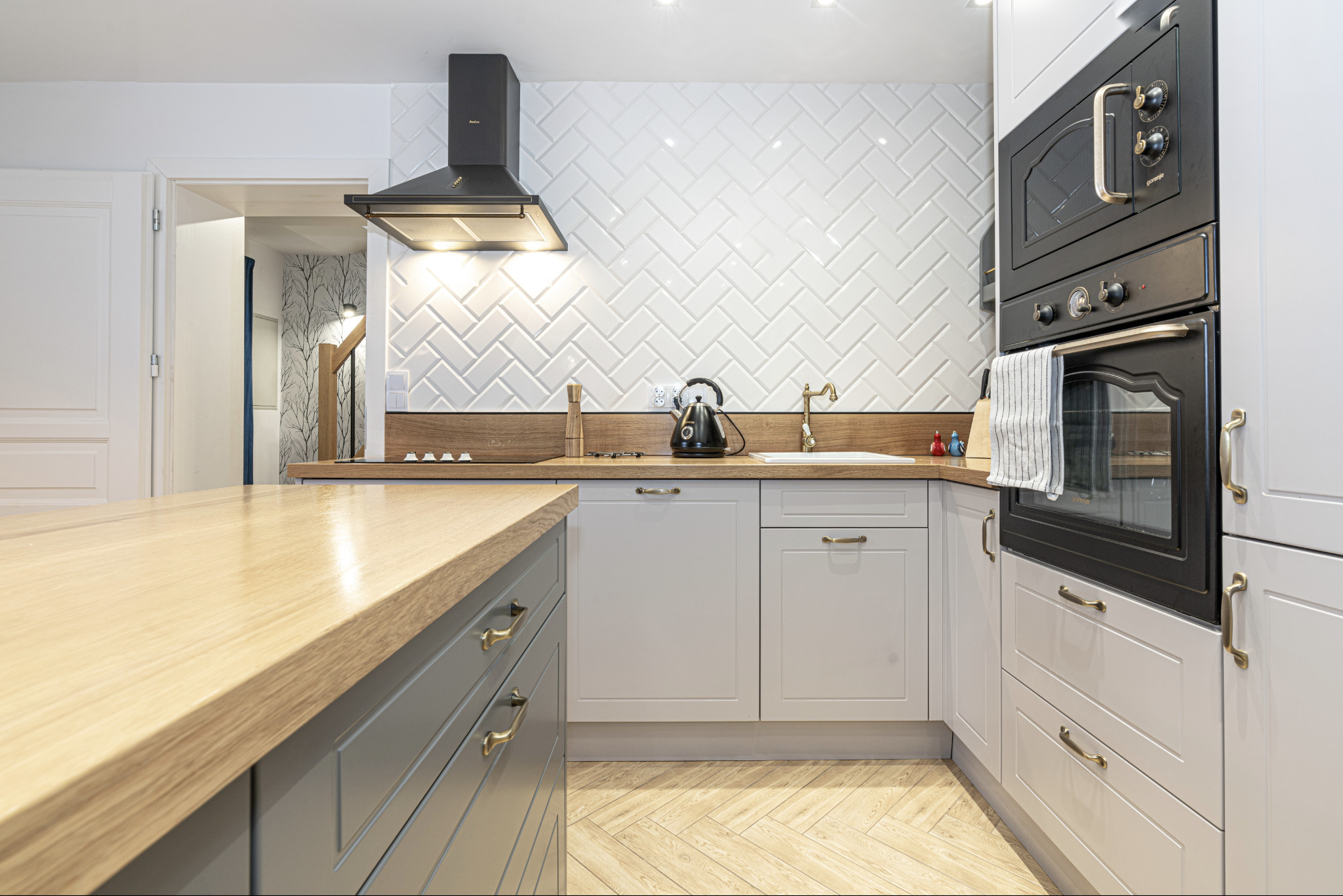 Jasna kuchnia z drewnianym blatem, białymi płytkami ułożonymi w jodełkę na ścianie, czarnym okapem i piekarnikiem oraz szarymi szafkami z mosiężnymi uchwytami, widok na otwarte drzwi do innego...