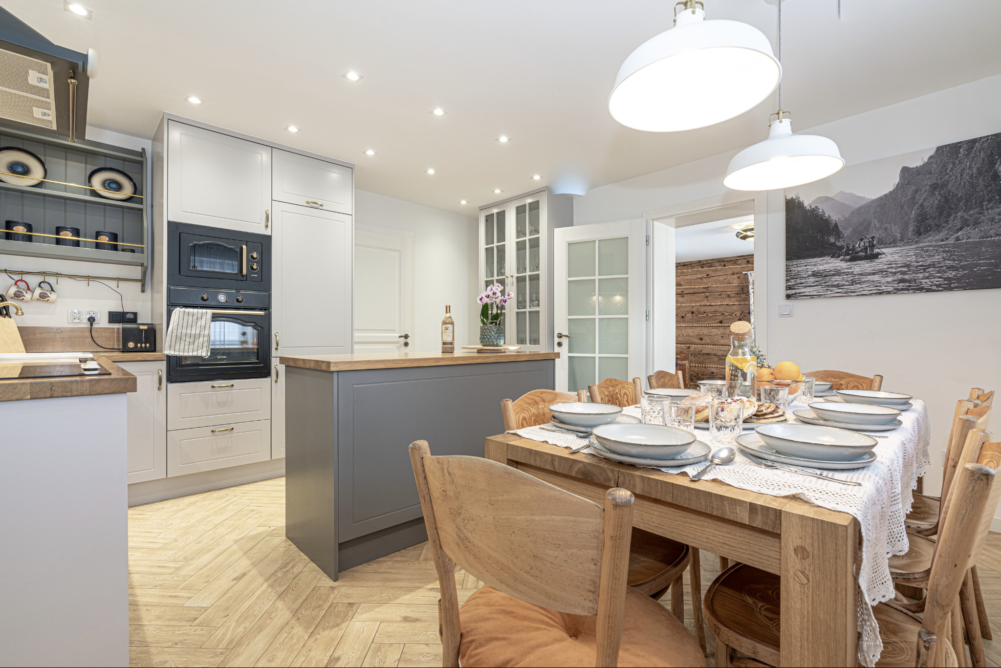 Jasna kuchnia z szarymi szafkami, drewnianym blatem i stołem z nakryciem na osiem osób, widok na ścianę z drewnianą boazerią w sąsiednim pomieszczeniu.
