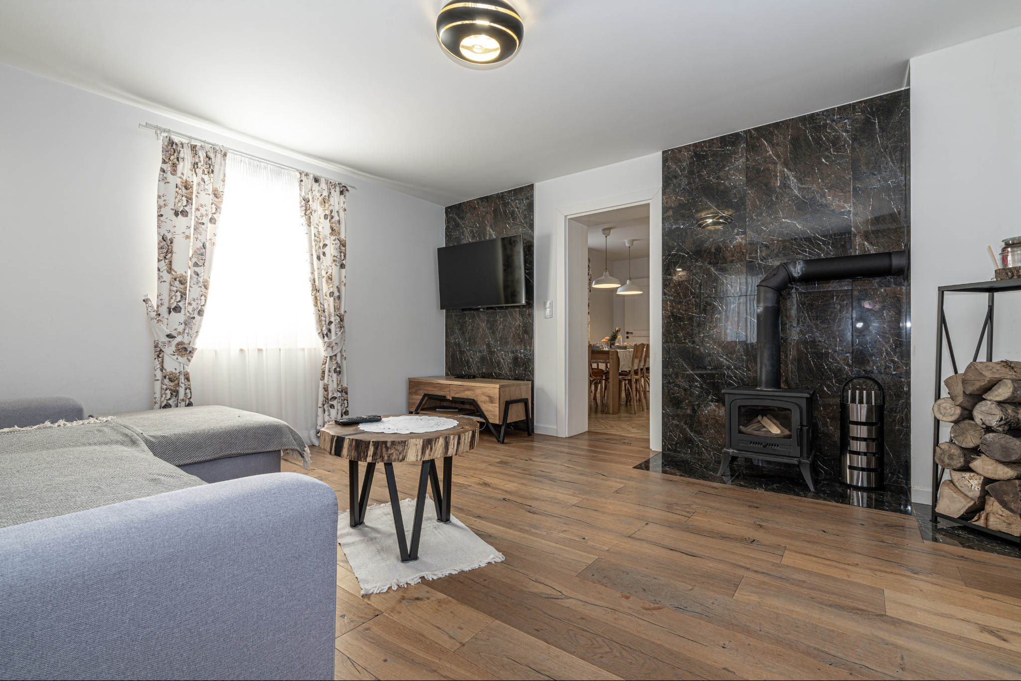 Salon z kominkiem obudowanym czarnym marmurem, drewnianą podłogą, stolikiem kawowym z plastra drewna i szarą sofą pod oknem z kwiecistymi zasłonami.