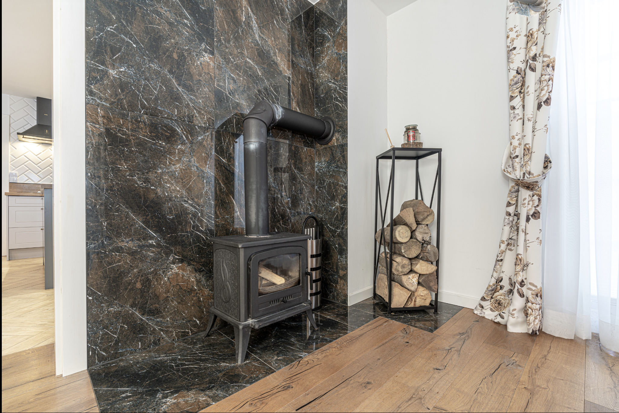 Rustykalny piec żeliwny z rurą dymową na tle marmurowej ściany, obok metalowy regał z ułożonym drewnem kominkowym, drewniana podłoga i zasłony w kwiaty.
