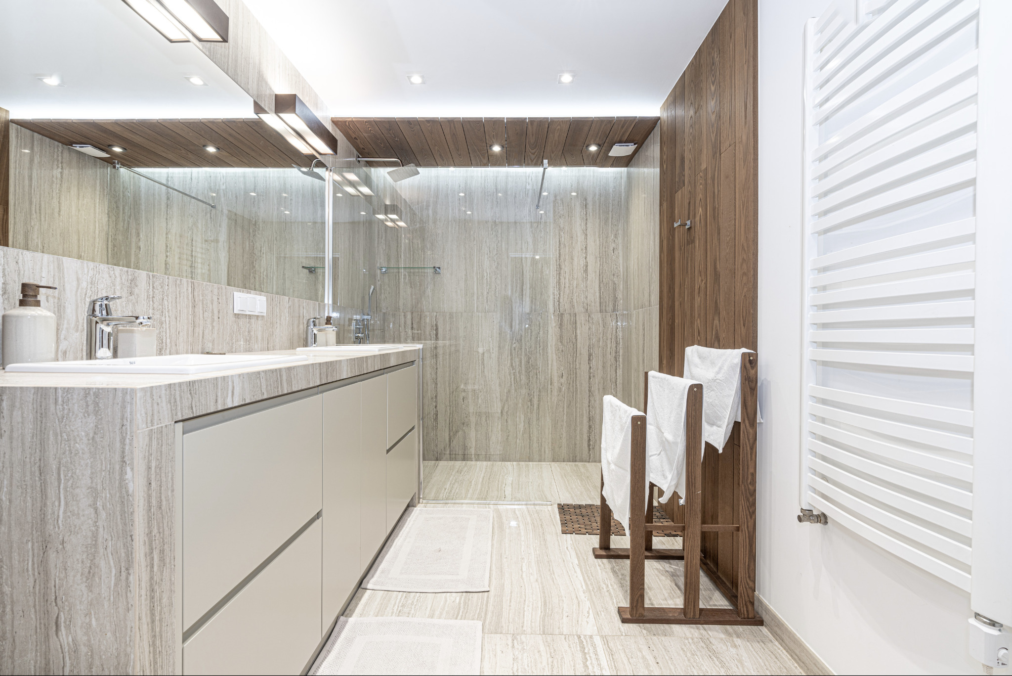 Nowoczesna łazienka z beżowymi płytkami imitującymi kamień, drewnianymi akcentami i przeszklonym prysznicem typu walk-in, uzupełniona o drewniany stojak na ręczniki i grzejnik drabinkowy.