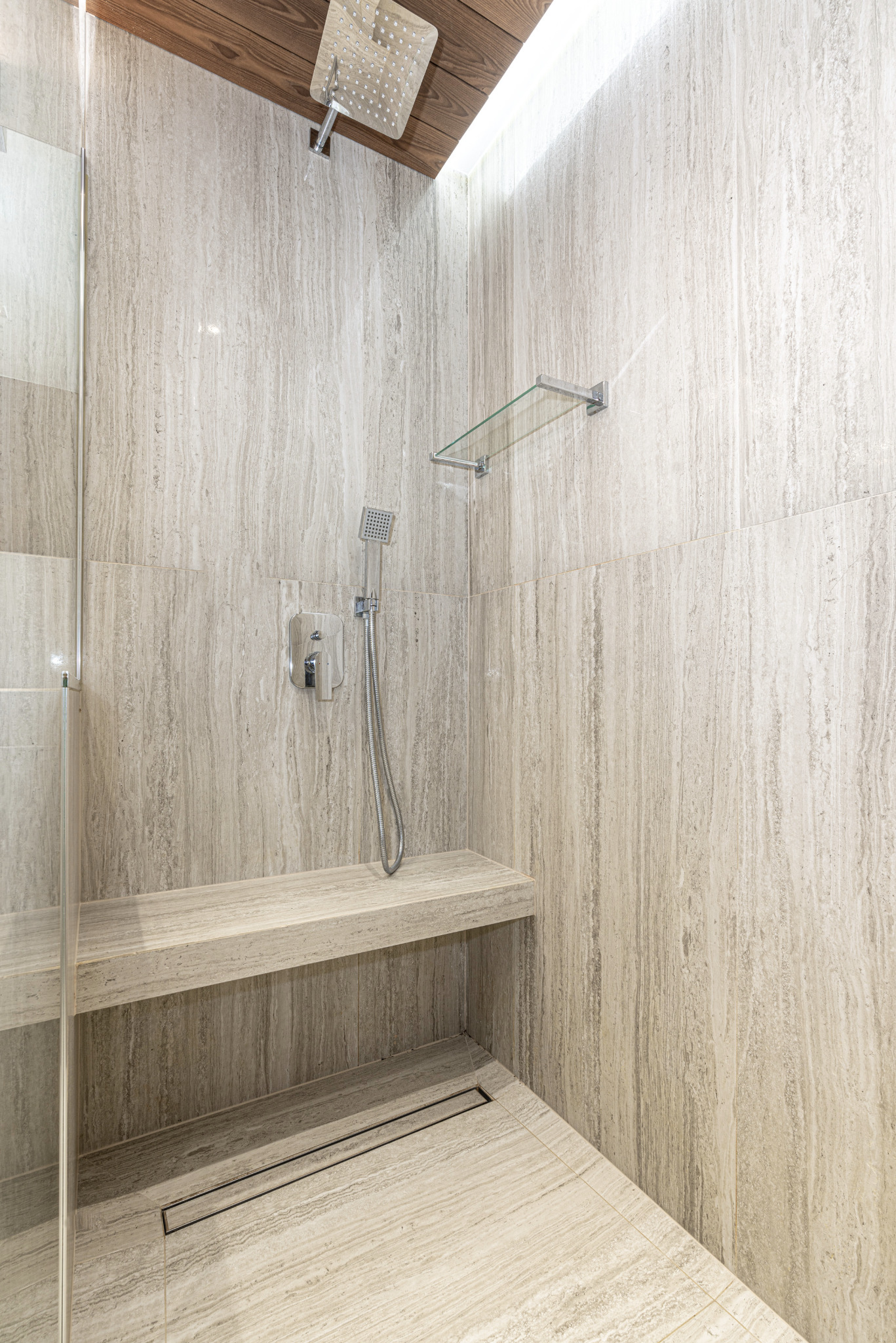 Nowoczesna kabina prysznicowa z siedziskiem i deszczownicą, wykończona płytkami imitującymi trawertyn, z odpływem liniowym w podłodze.