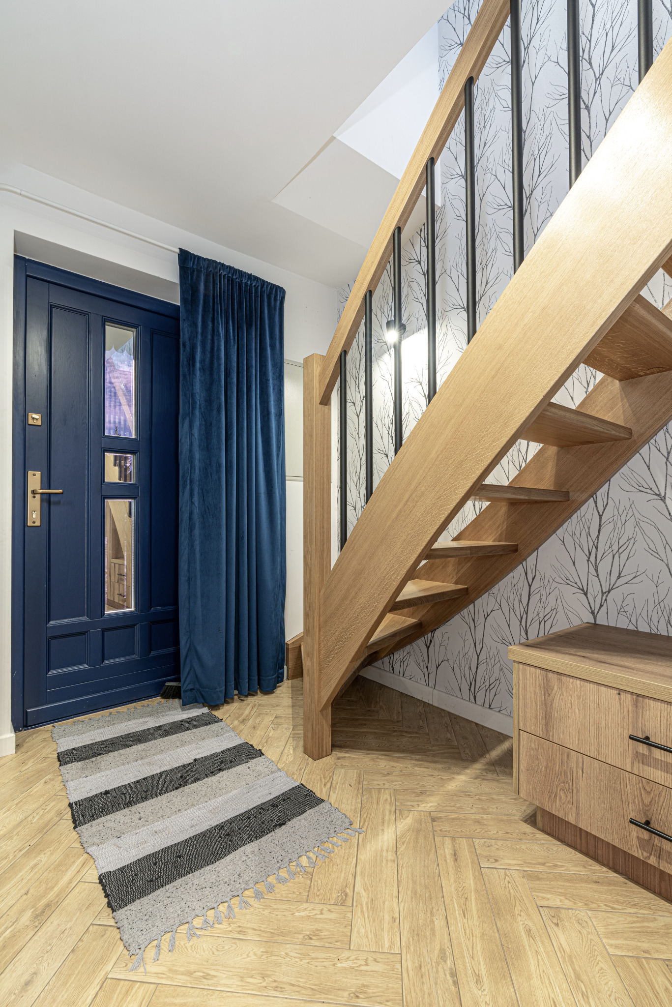 Nowoczesny przedpokój z drewnianymi schodami z czarnymi balustradami, niebieskie drzwi wejściowe z aksamitną zasłoną, dywanik w paski i komoda z szufladami na parkiecie w jodełkę.