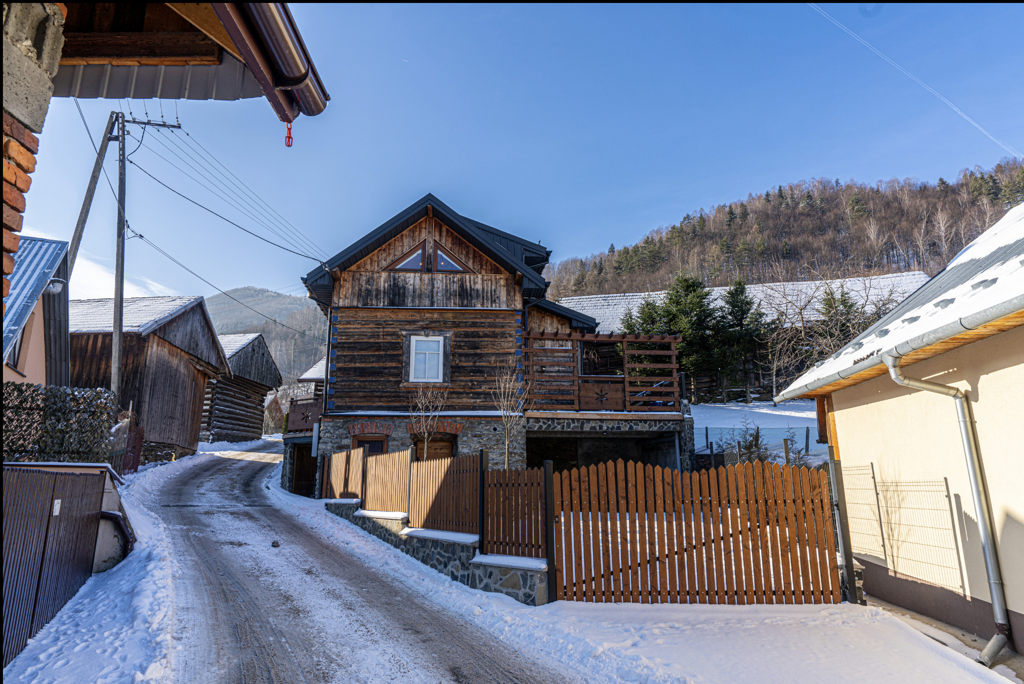 Drewniany dom z bali z podmurówką z kamienia i brązowym płotem w zimowej scenerii, widok z dołu drogi pokrytej śniegiem.