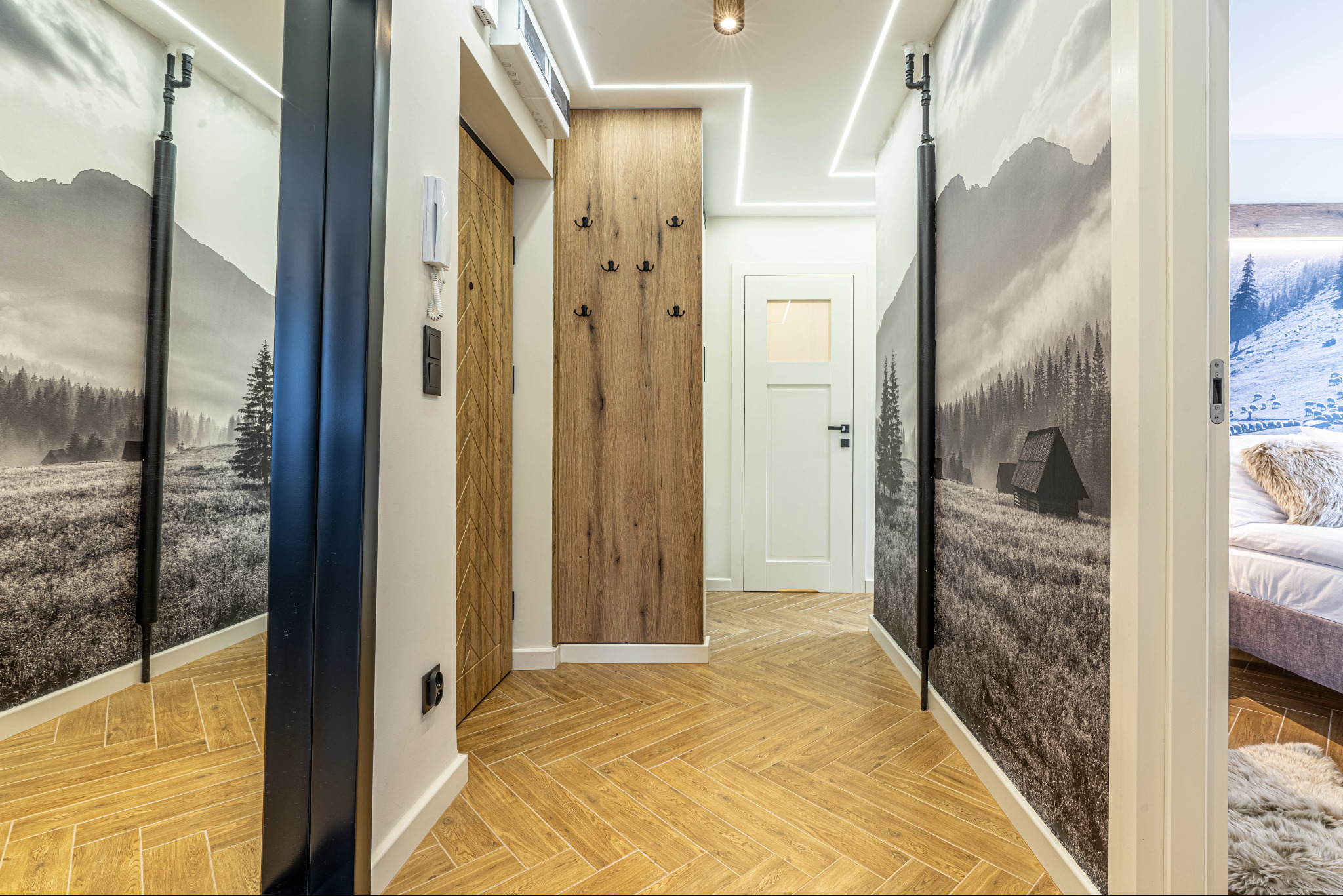 Wnętrze korytarza z podłogą ułożoną w jodełkę, drewnianym panelem z wieszakami, lustrem w czarnej ramie i fototapetą z motywem górskim.