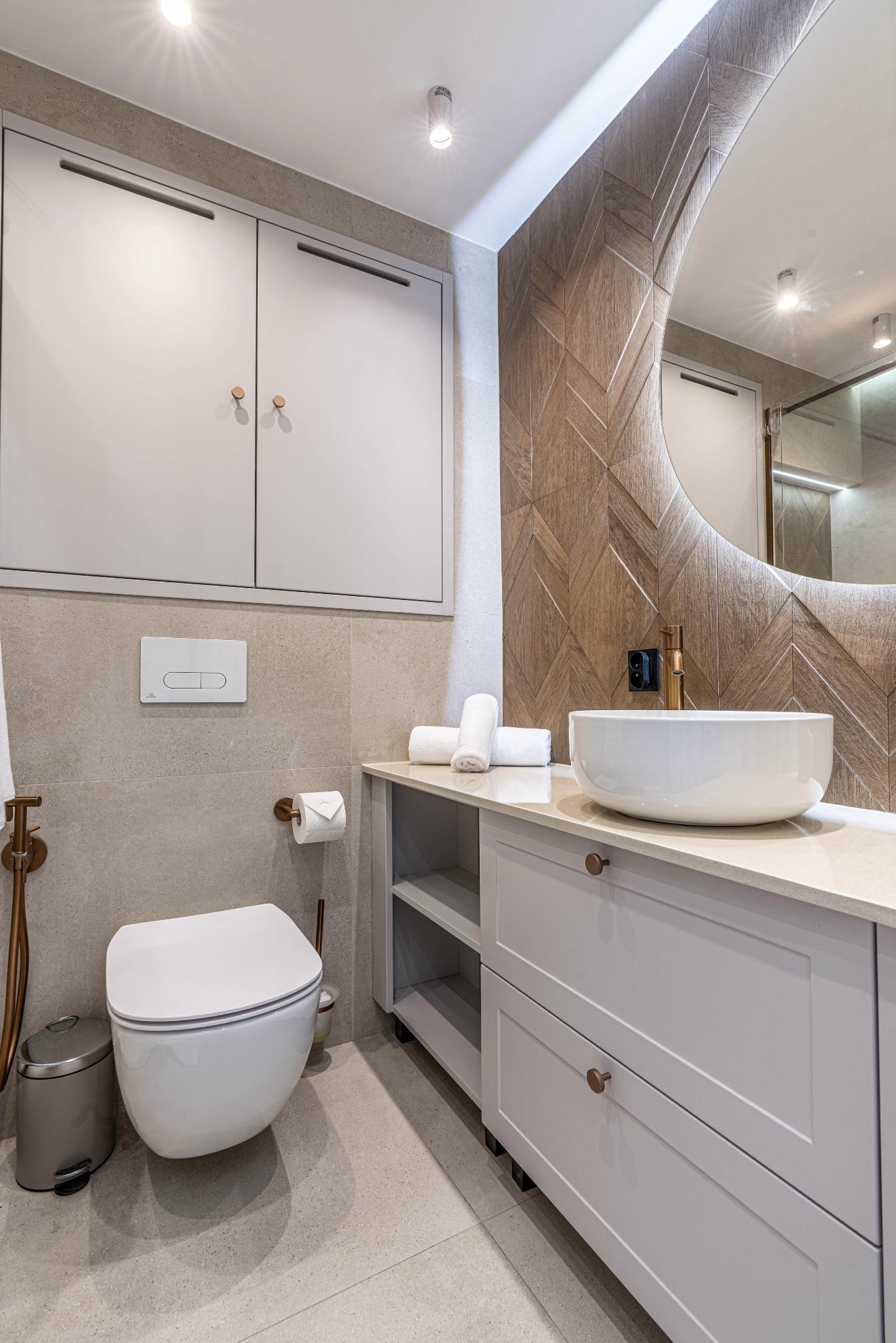 Nowoczesna łazienka z podwieszaną toaletą, szafką z umywalką nablatową, okrągłym lustrem z podświetleniem i drewnianą mozaiką na ścianie