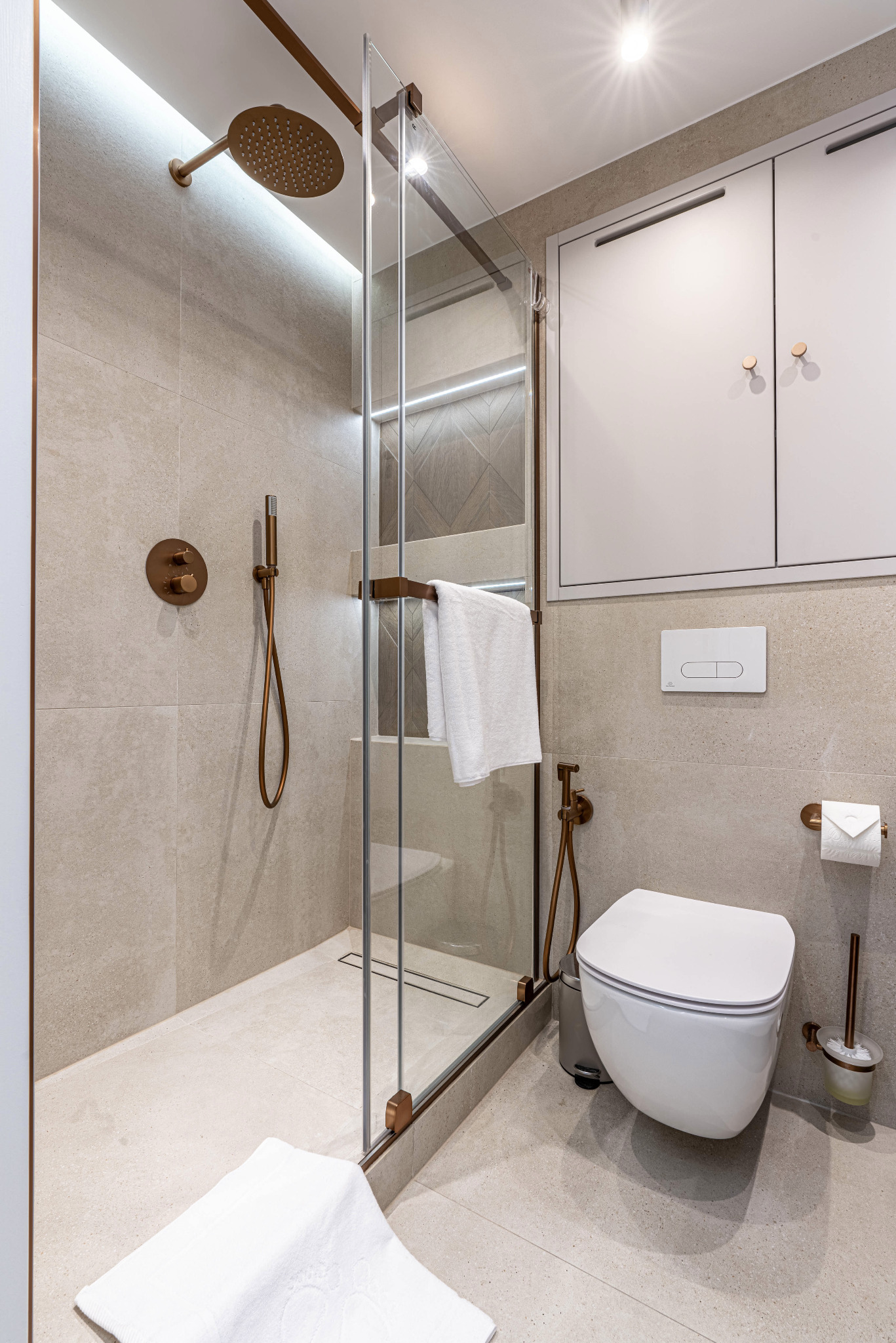 Nowoczesna łazienka z kabiną prysznicową typu walk-in, armaturą w kolorze miedzi i wiszącą toaletą, wykończona płytkami w odcieniach beżu.