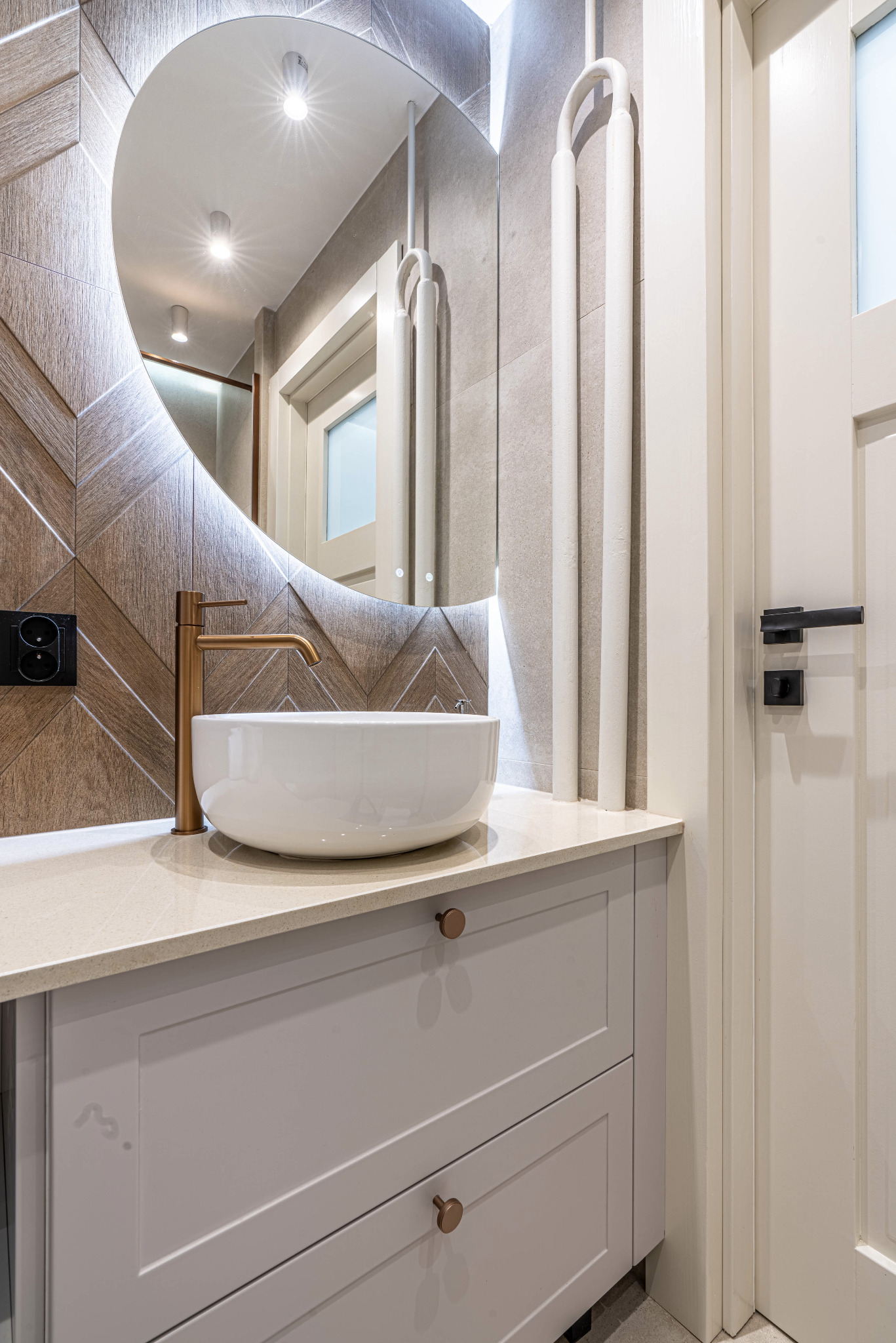 Nowoczesna łazienka z okrągłym, podświetlanym lustrem, białą umywalką nablatową, złotą baterią, drewnianą ścianą w jodełkę i białymi grzejnikami dekoracyjnymi.