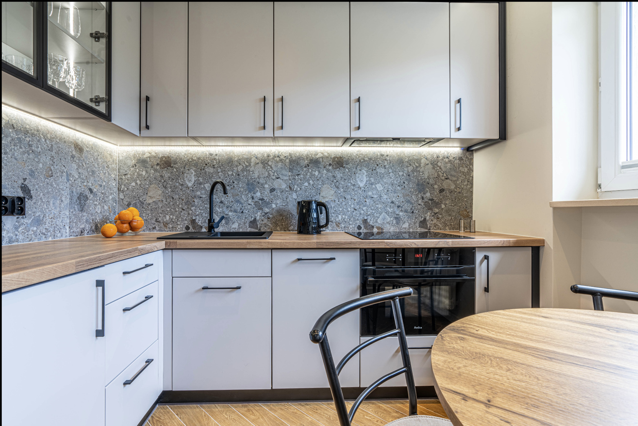 Jasna kuchnia z szarym kamiennym backsplashiem, czarną armaturą i drewnianym blatem, oświetlona listwami LED pod szafkami, z widocznym fragmentem okrągłego stołu i krzesła