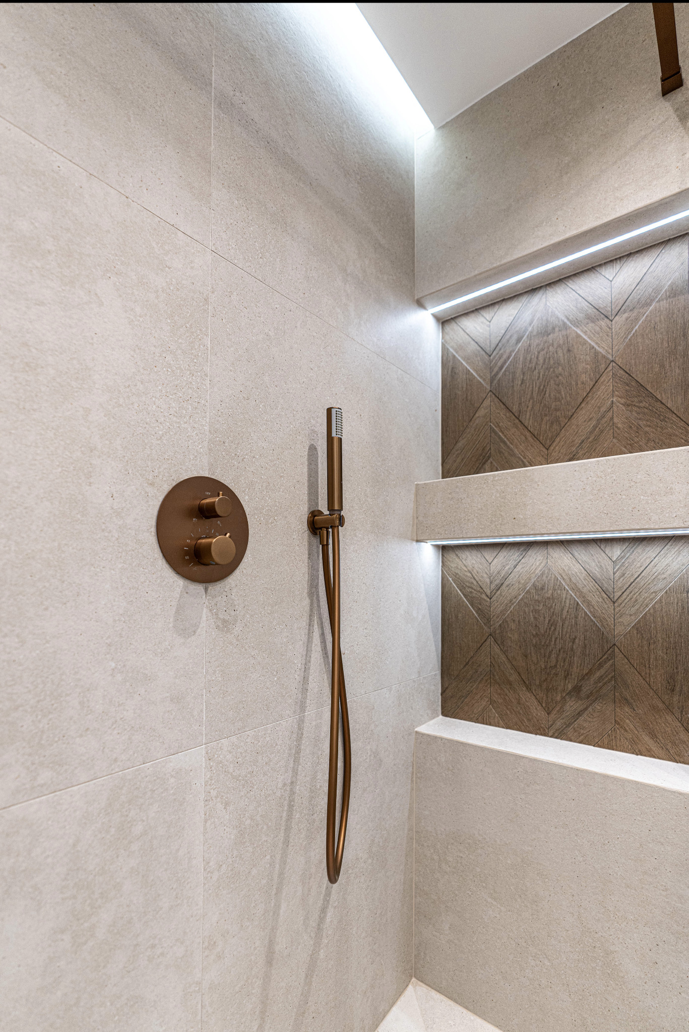 Nowoczesna kabina prysznicowa z beżowymi płytkami, dekoracyjną ścianą z drewnianym wzorem chevron, miedzianą armaturą i subtelnym oświetleniem LED.