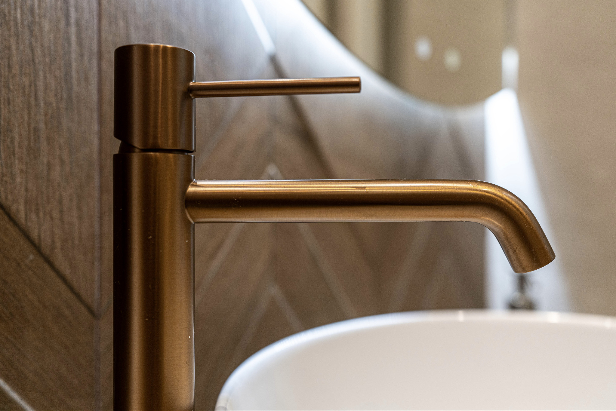 Miedziana bateria umywalkowa z prostą wylewką i uchwytem, na tle ściany z wzorem w jodełkę i fragmentu białej umywalki.