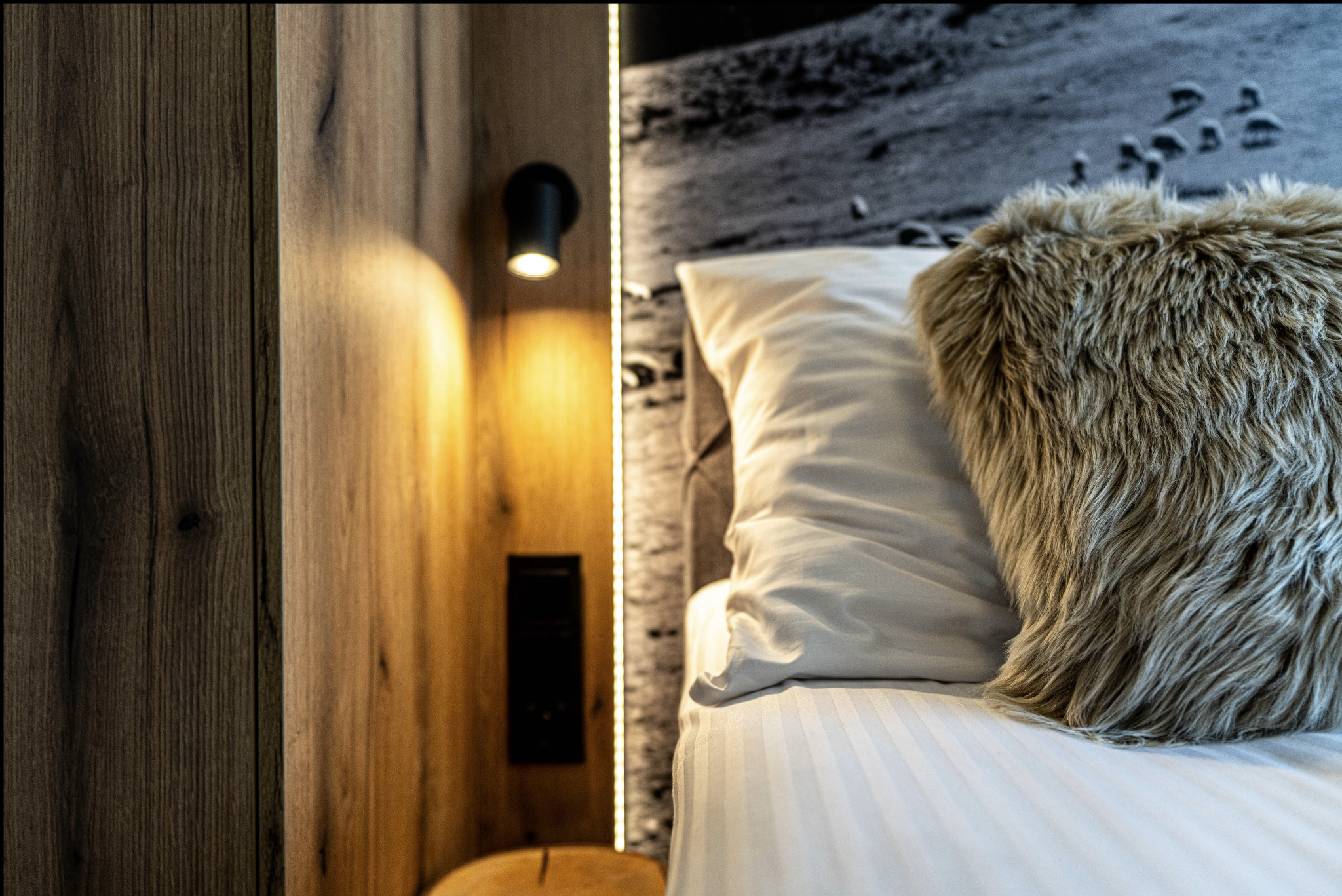 Nowoczesna sypialnia z drewnianą ścianą, oświetleniem punktowym i dekoracyjną poduszką z długim włosiem na łóżku z białą pościelą.