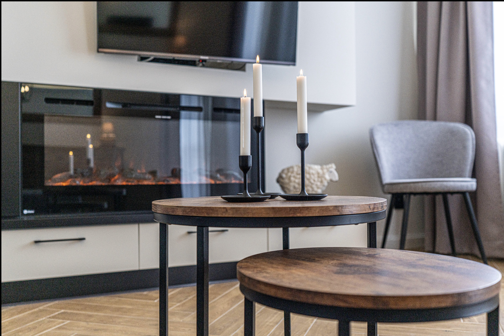 Nowoczesny salon z elektrycznym kominkiem, stolikami kawowymi w stylu industrialnym, trzema świecami w czarnych świecznikach i dekoracyjną figurką owcy.