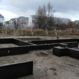 Wykopane i zalane fundamenty pod budynek mieszkalny na działce budowlanej, z widocznymi budynkami mieszkalnymi w tle.