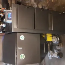 Nowoczesny, dwuczęściowy piec z widocznym panelem sterowania, rurami i elementami instalacji hydraulicznymi na tle betonowej ściany.