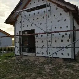 Budynek w trakcie ocieplania styropianem, widoczne rusztowanie, nieotynkowana elewacja z przyklejonymi płytami styropianowymi i zaślepkami, fragment dachu z dachówką.
