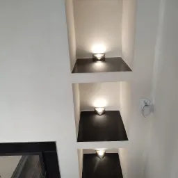 Wnęki ścienne z czarnymi półkami i trójkątnymi lampami, obok czarny wkład kominkowy, widoczny fragment białej ściany z gniazdkiem.