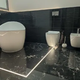 Nowoczesna łazienka z czarnymi płytkami ściennymi o falistej strukturze, wolnostojąca wanna z chromowaną armaturą, podwieszana toaleta i bidet, podświetlana listwa przypodłogowa na posadzce...