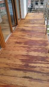 Drewniana podłoga na balkonie z widocznymi słojami i przebarwieniami, balustrada ze stali nierdzewnej w tle.