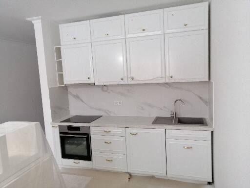 Nowoczesna, biała kuchnia z marmurowym blatem i backsplashiem, widoczny piekarnik i zlewozmywak z baterią, górne szafki z delikatnymi uchwytami.