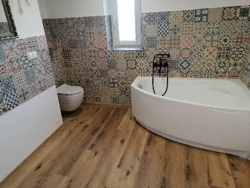 Wykończona łazienka z drewnopodobną podłogą, patchworkowymi płytkami na ścianach, białą wanną narożną i toaletą.