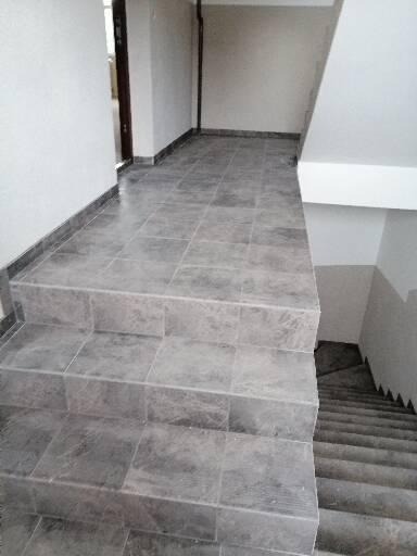 Wykończone schody wewnątrz budynku, pokryte szarymi płytkami ceramicznymi imitującymi kamień, widoczne fugi i krawędzie stopni, perspektywa z góry.