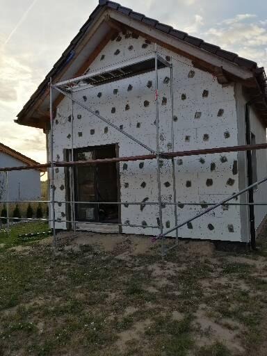 Budynek w trakcie ocieplania styropianem, widoczne rusztowanie, nieotynkowana elewacja z przyklejonymi płytami styropianowymi i zaślepkami, fragment dachu z dachówką.
