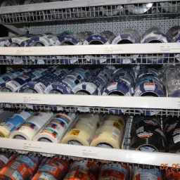 Półki regału w składzie budowlanym wypełnione różnymi rodzajami taśm malarskich i budowlanych marki Blue Dolphin, w tym taśmy do folii izolacyjnej, taśmy do stolarki budowlanej oraz taśmy Duct Tape...