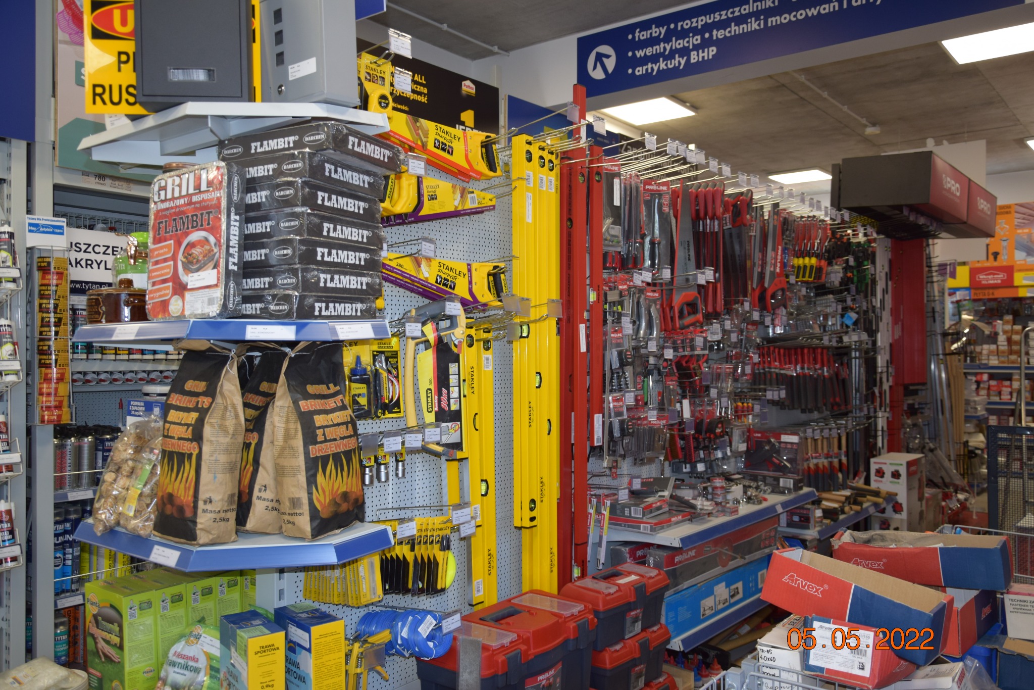 Wnętrze sklepu budowlanego: półki z narzędziami Stanley, poziomice, węgiel drzewny do grilla, zaprawy murarskie, farby i artykuły BHP.