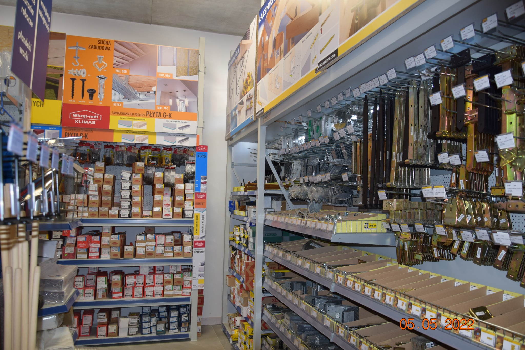 Wnętrze sklepu budowlanego z regałami wypełnionymi różnorodnymi materiałami i akcesoriami montażowymi, w tym mocowania do płyt gipsowo-kartonowych, widoczne reklamy producentów na półkach.